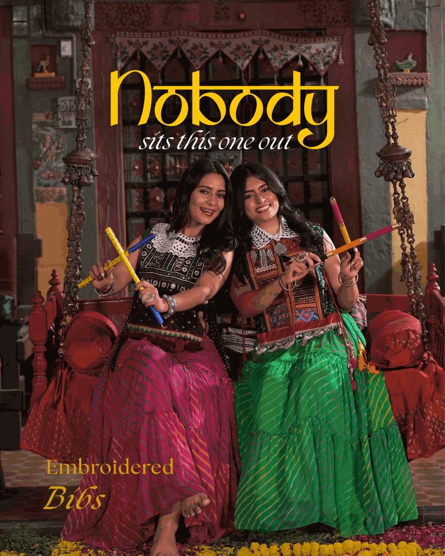Two women in embroidered bibs and tiered lehariya skirts, one pink and one green, sit together on a traditional wooden swing in an old-style house, each holding dandiya sticks. The headline reads Nobody sits this one out.